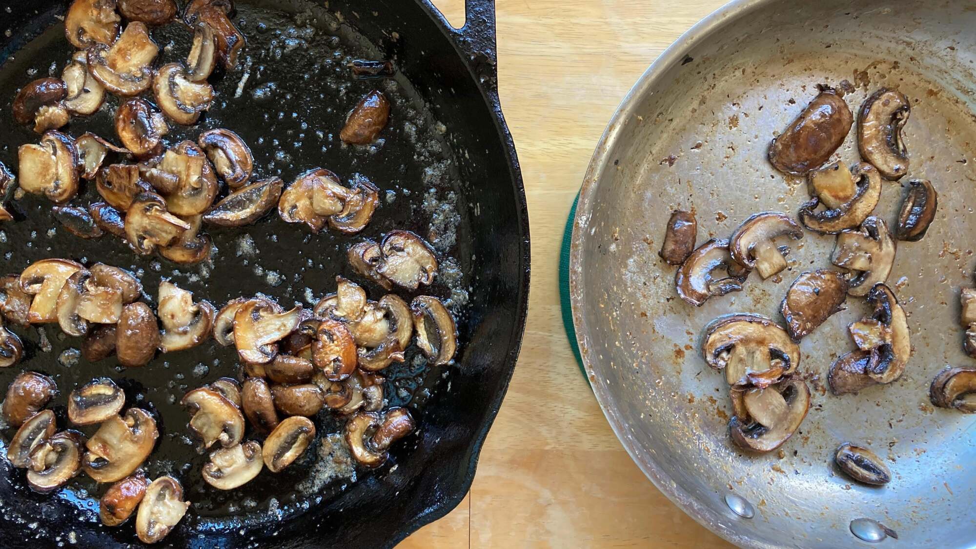 Top-down photo of two pans of sautéed sliced mushrooms, side by side on a light wood table. On the left side is a large black cast iron skillet and on the right is a smaller stainless steel skillet.