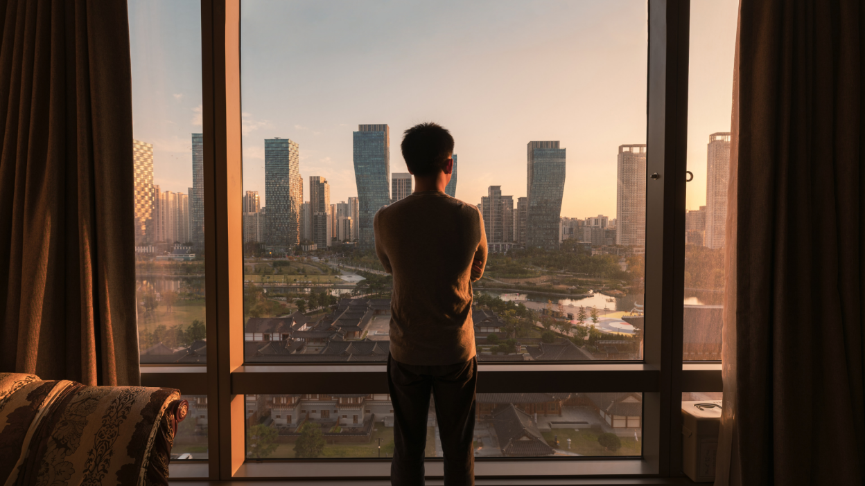 Man inside a high rise apartment standing in front of a large set of windows, looking out onto the city at sunset